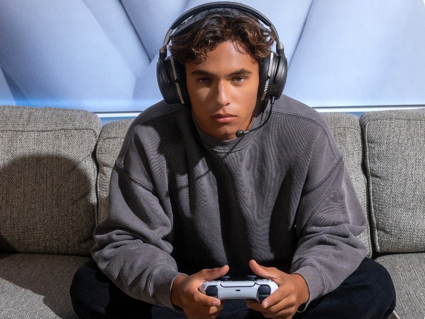 Headset Audeze Maxwell 2 ANC - PlayStation čierny Hráč sediaci na pohovke s ovládačom v rukách a headsetom Audeze Maxwell 2 ANC na hlave