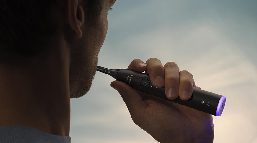 Muž používa elektrickú zubnú kefku Philips Sonicare 6500 HX7411/10 pri čistení zubov, drží čiernu kefku s podsvietenou rukoväťou.