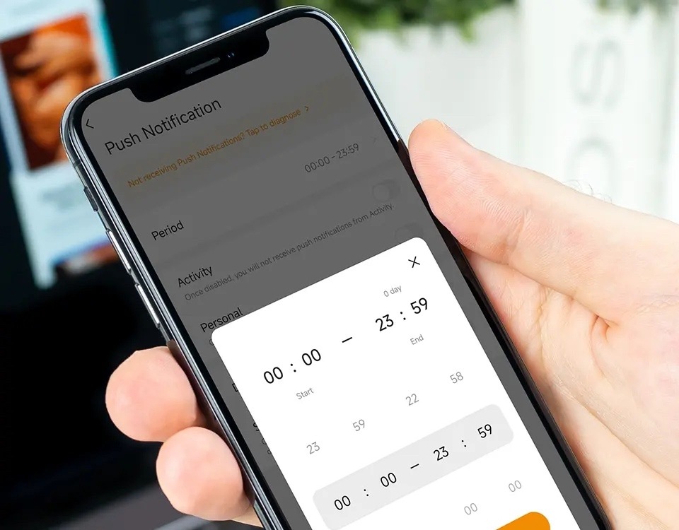 Na obrázku je ruka držiaca chytrý telefón s otvorenou aplikáciou Imou. Na displeji je nastavenie push notifikácií, kde je možné určiť časové obdobie pre prijímanie alebo vypnutie upozornenia z kamery. Uprostred obrazovky je otvorené okno s nastavením času od 00:00 do 23:59 pomocou posuvných číselných hodnôt. Obrázok ukazuje možnosť prispôsobenia oznámenia podľa dennej doby.