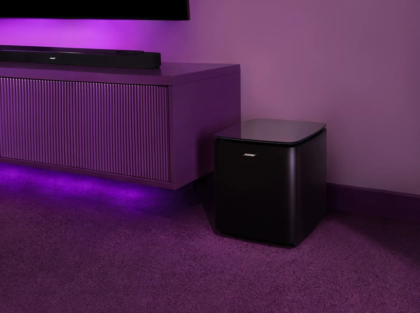 Subwoofer Bose Lifestyle Ultra čierny Subwoofer Bose Bass Module umiestnený na podlahe pri TV stolíku s fialovým podsvietením.