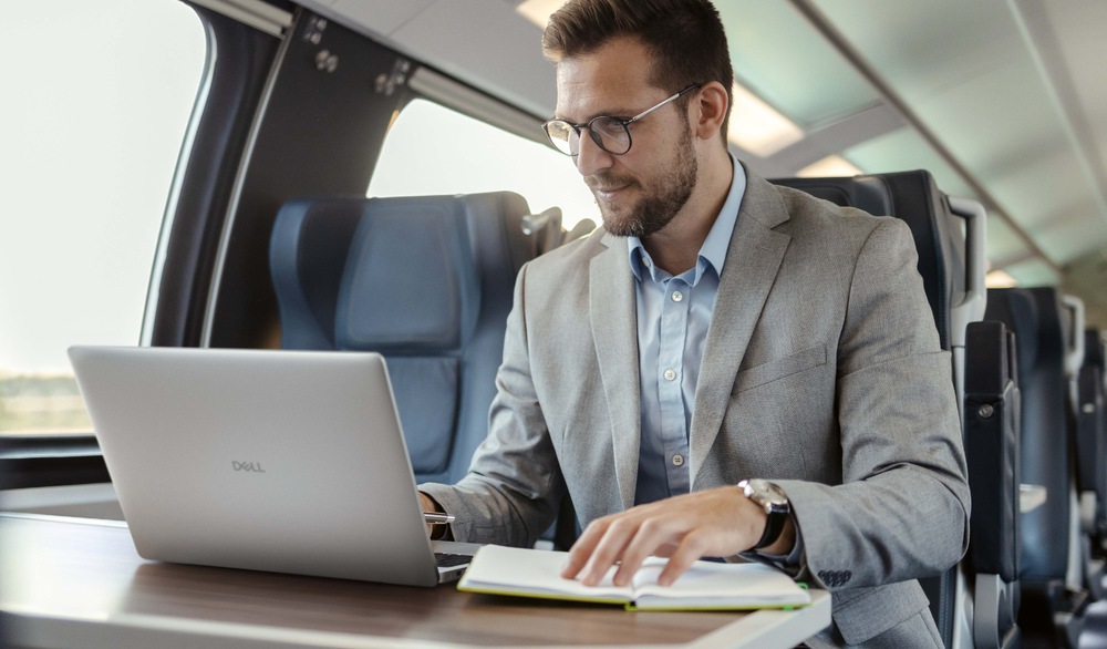 Notebook Dell Pro 16 PC16255 sivý Muž v obleku pracuje na notebooku pri stole vo vlaku. Vyzerá sústredene, má otvorené dokumenty. Interiér vlaku je moderný a dobre osvetlený.