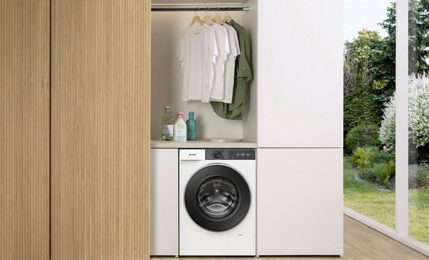 práčka Gorenje W484A3 UltraClean vedľa drevenej šatníkovej skrine. Nad práčkou visí na ramienkach oblečenia. Na pravej strane sú veľké presklené dvere do záhrady.