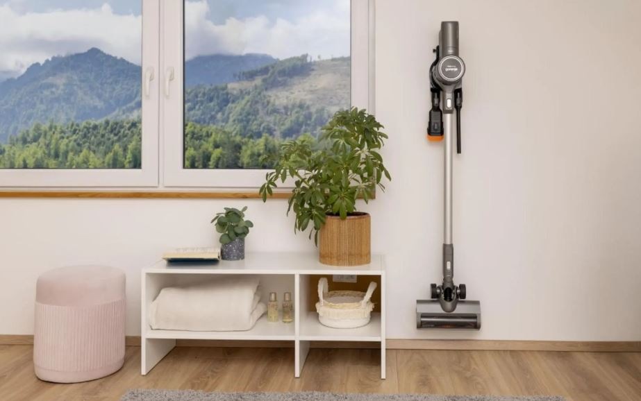 Tyčový vysávač Gorenje SVC252420FFA FreeForce InverterPower sivý tyčový vysávač Gorenje SVC252420FFA visi na stene vo svojom držiaku. Vedľa je okno a pod ním policova skrinka.