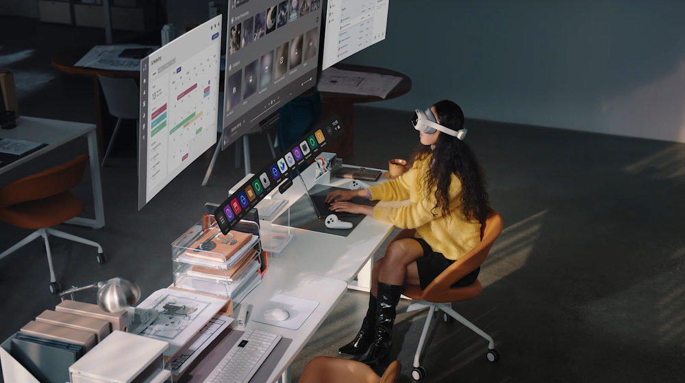 Okuliare pre virtuálnu realitu PICO 4 Ultra biele Žena v žltom svetre s nasadeným VR headsetom sedí pri modernom stole a interaguje s plávajúcimi digitálnymi obrazovkami. Scéna evokuje futuristický pracovný priestor.