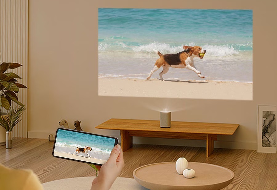 Laser TV Samsung SP-LPF5DSA The Premiere 5 premieta na stenu záber psa bežiaceho po pláži, v popredí ľudská ruka drží mobilný telefón, na ktorého obrazovke je rovnaký záber
