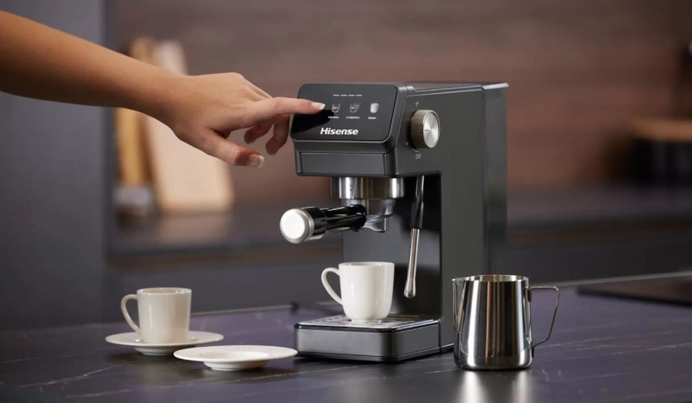 Espresso Hisense HESCM20LP antracitová farba ženská ruka vyberá program na pákovom kávovare Hisense HESCM20LP. Vedľa kávovaru sú hrnčeky a nádobka na šľahanie mlieka.