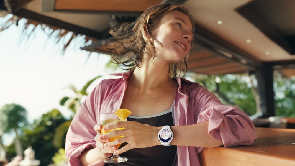 Inteligentné hodinky Huawei Watch 6 Pro 46 mm čierne/titánová farba Usmievajúca sa žena s mokrými vlasmi odpočíva pri bare s nápojom v ruke; na zápästí má inteligentné hodinky s elegantným analógovým ciferníkom