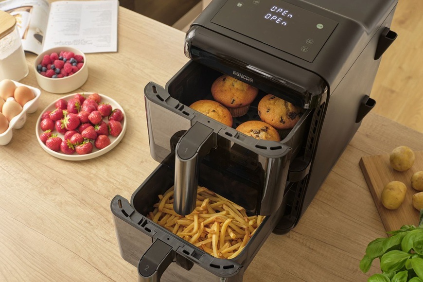 Fritéza teplovzdušná Sencor SFR 1250BK čierna Teplovzdušná fritéza Sencor SFR 1250BK s vysunutými dvoma košmi ukazuje súčasnú prípravu rôznych pokrmov, kedy v spodnom koši sú hranolky av hornom muffiny. Na kuchynskej linke sú pripravené čerstvé suroviny ako jahody, vajcia a zemiaky.