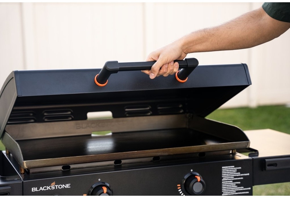 Ruka zdvíhajúca pokrievku grilu Blackstone za ergonomické plastové madlo s oranžovými dorazmi.