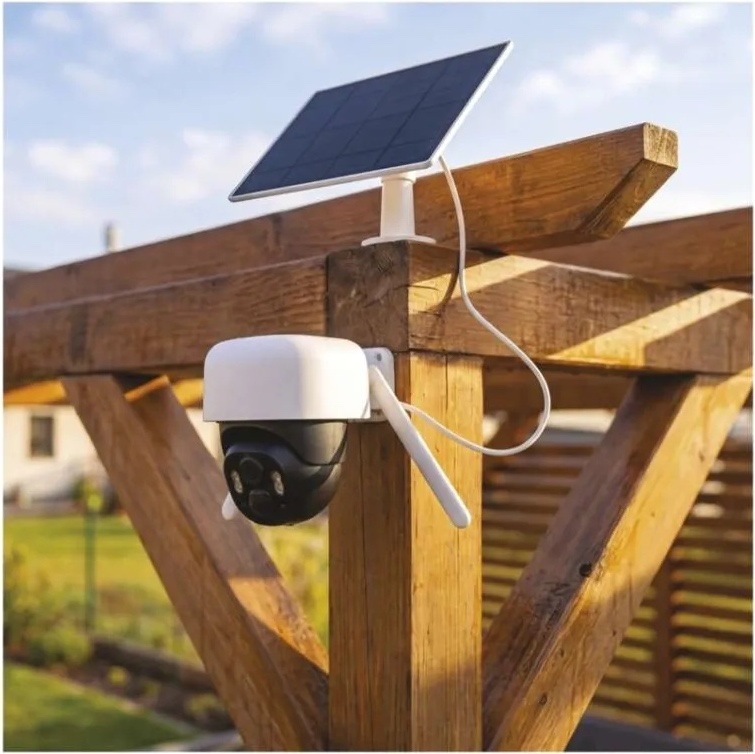 IP kamera EMOS GOSMART venkovní, Wi-Fi IP-1200 WASP 5MPX biela Otočná kamera EMOS GoSmart je namontovaná na drevenom stĺpiku pergoly. Nad ňou je solárny panel v bielom ráme, prepojený s kamerou káblom na dobíjanie.