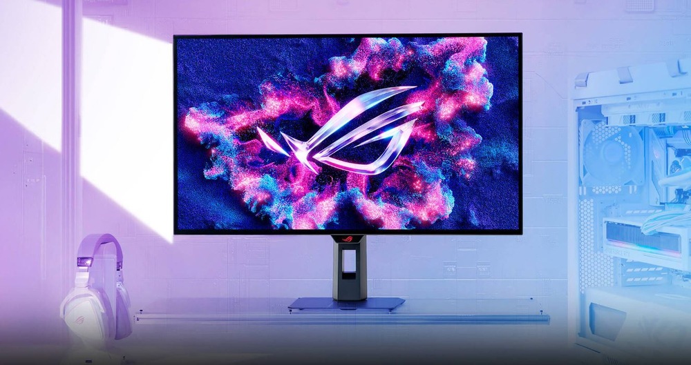 Elegantný herný monitor zobrazuje na obrazovke žiarivé, futuristické logo obklopené farebnou hmlistou grafikou. Vedľa neho ležia biele slúchadlá a rozsvietený herný počítač, ktoré navodzujú modernú atmosféru technickej vyspelosti.