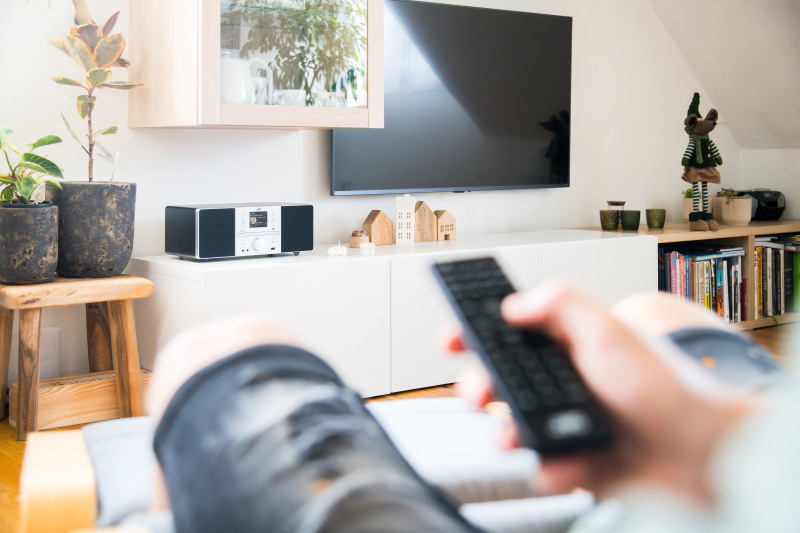 Moderná domáca obývacia izba s mikrosystémom JVC RD-E874B umiestneným na bielej skrinke pod televíziou, fotená z pohľadu osoby držiacej diaľkový ovládač