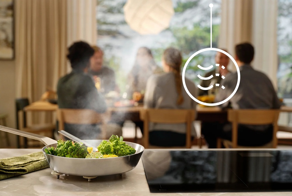 V popredí je indukcia Electrolux. Vedľa je položená panvica so zeleninou. V pozadí sedia ľudia pri stole. Nad doskou je symbol pre funkciu Breeze.