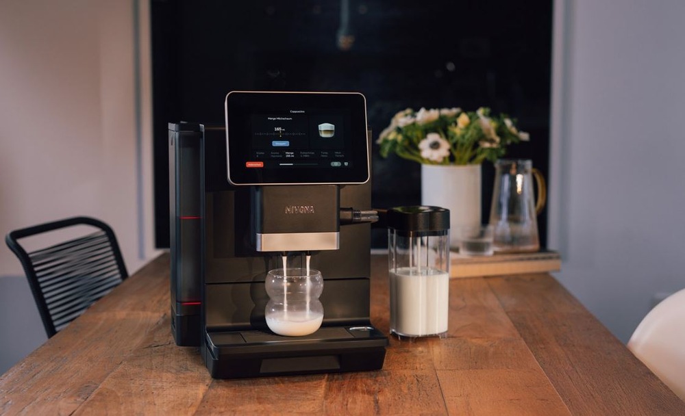 Kávovar Nivona stojí na kuchynskom stole a pripravuje mliečny nápoj. Na displeji je vidieť prubeh pripravy nápoja. Za kávovarom je váza s kyticou a džbán s vodou
