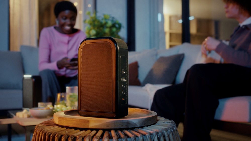 Soundbar JBL BAR 1300 MK2 čierny Dvaja ľudia sa rozprávajú v obývacej izbe, žena drží mobilný telefón, v popredí je odnímateľný bezdrôtový reproduktor soundbaru JBL BAR 1300 MK2 na stolíku