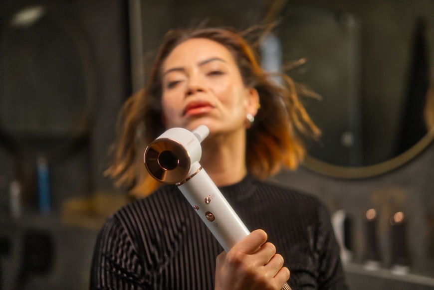 Žena s krátkymi vlasmi používa fúkaciu kulmu Sencor SHS 9100RS Artemiss, ktorú drží v ruke pri úprave účesu. Prúd vzduchu z kulmy dvíha pramene vlasov, zatiaľ čo pozadie tvorí interiér kúpeľne so zrkadlom a kozmetickými doplnkami.