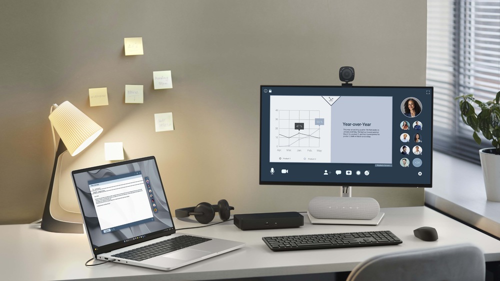 Notebook Dell Pro 16 PC16255 sivý Moderný pracovný stôl s notebookom a na doske stola umiestneným monitorom, na ktorom sa zobrazujú grafy a rozhrania pre videohovory. Na stene sú nalepené poznámkové lístky, lampička a zelené rastliny dodávajú miestnosti útulný vzhľad.
