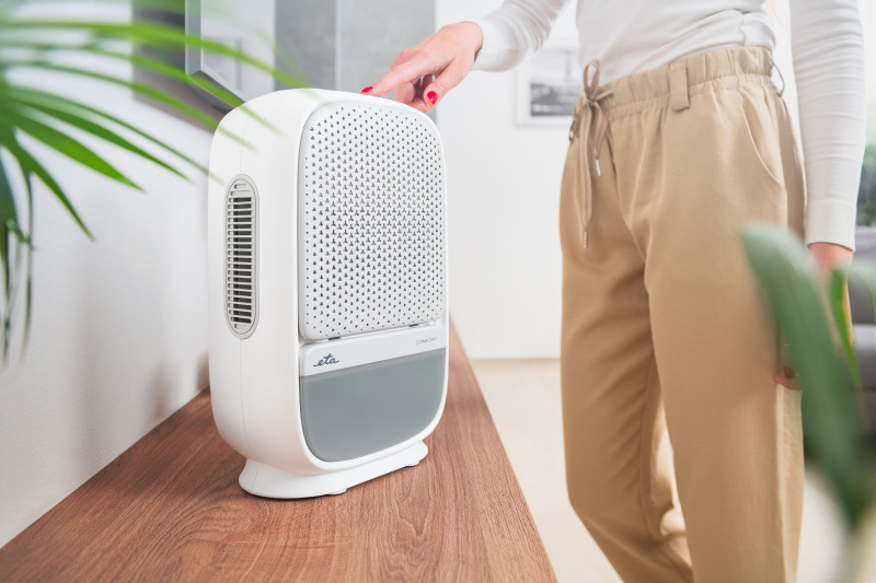 Modermí bielo-sivý odvlhčovač vzduchu ETA Comfort s perforovanou čelnou doskou a integrovanou rukoväťou, ktorý je ovládaný dotykovo ženskou rukou