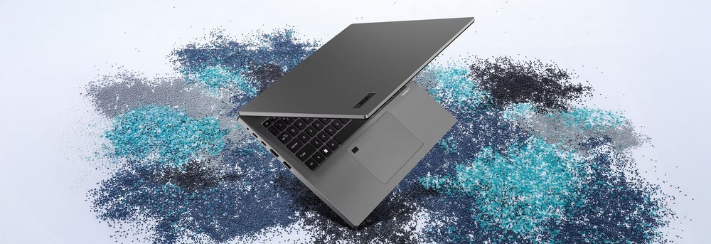 Notebook Acer TravelMate P2 14 (TMP214-56-TCO-32RF) strieborný Elegantný šedý notebook, mierne otvorený, leží na štruktúrovanom povrchu s rozptýlenými modrými a šedými časticami.