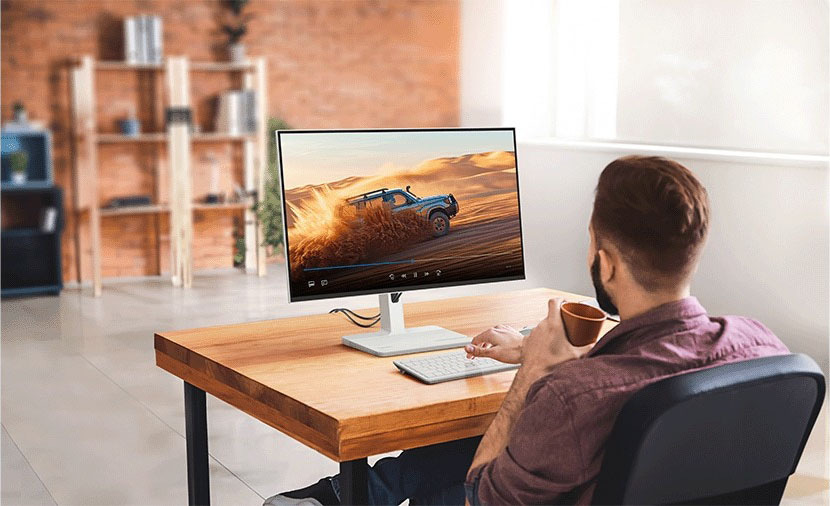Monitor Lenovo L27h-4A sivý Muž sedí pri drevenom stole v modernej kancelárii, drží hrnček s kávou a na obrazovke počítača sleduje terénne vozidlo rútiace sa pieskom.