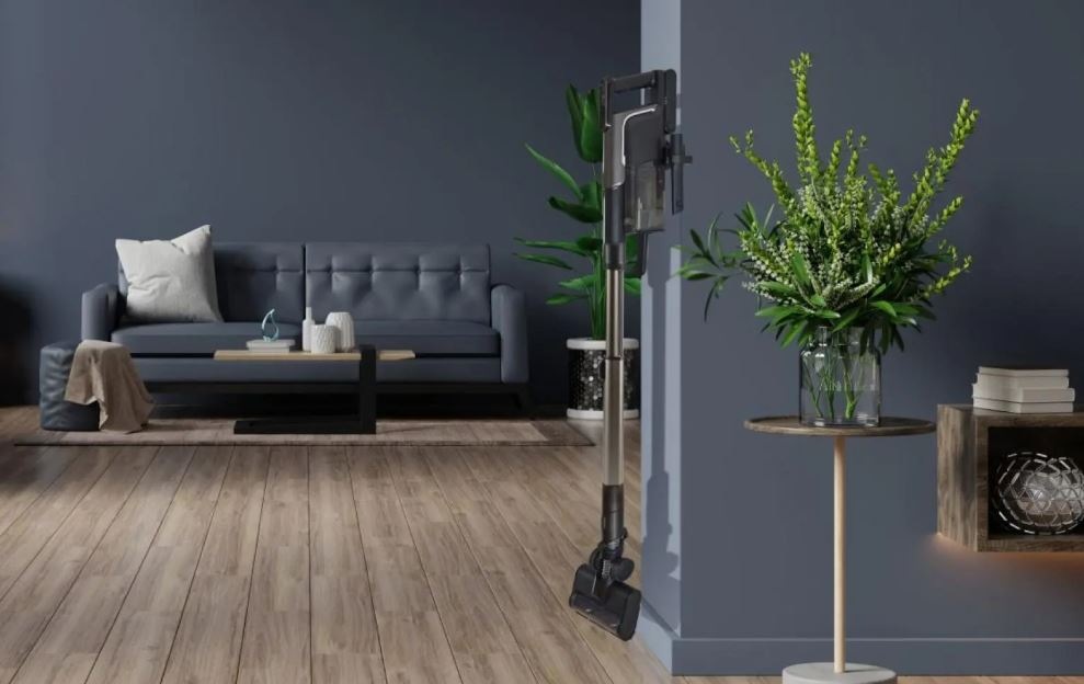 Tyčový vysávač Hisense HVC646630A Hi-Move VI Adapt InverterTech čierny vysávač Hisense HVC646630A visí v stanici na stene v izbe s tmavými stenami a vybavením.. Vedľa vysávača je rastlina.