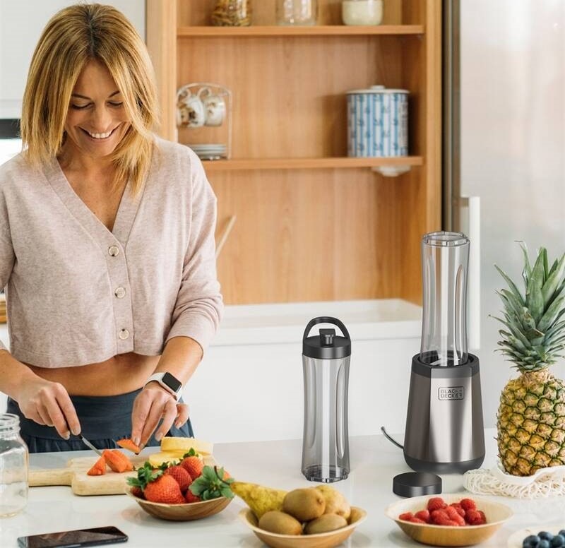 Usmievavá žena pripravuje na kuchynskej linke ovocie pre čerstvé smoothie, vedľa toho je umiestnený mixér Black+Decker BXJB1204E