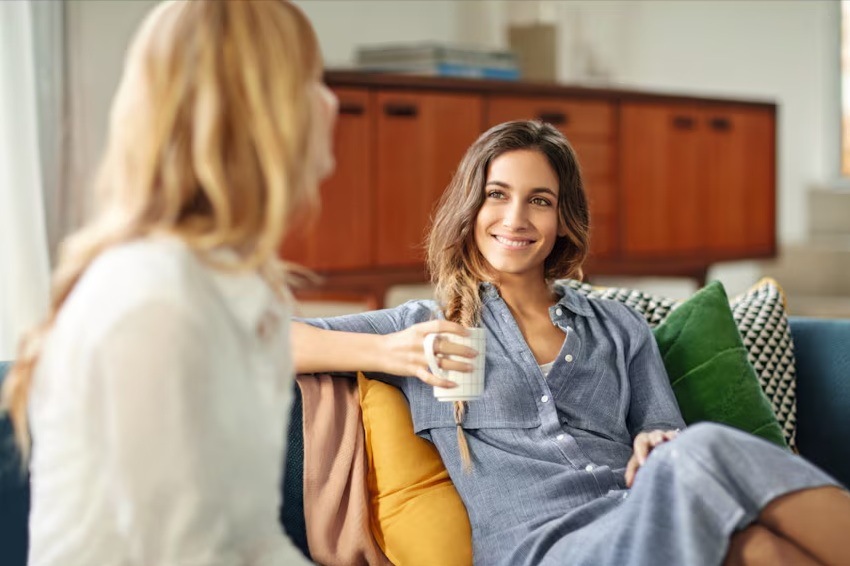 Práčka Electrolux Série 600 EW6F8482C biela Dve ženy sedia na pohovke v obývačke a konverzujú. Jedna je chrbtom a druhá čelom. Usmieva sa a drží šálku. Opiera sa o farebné vankúše. V pozadí je komoda.