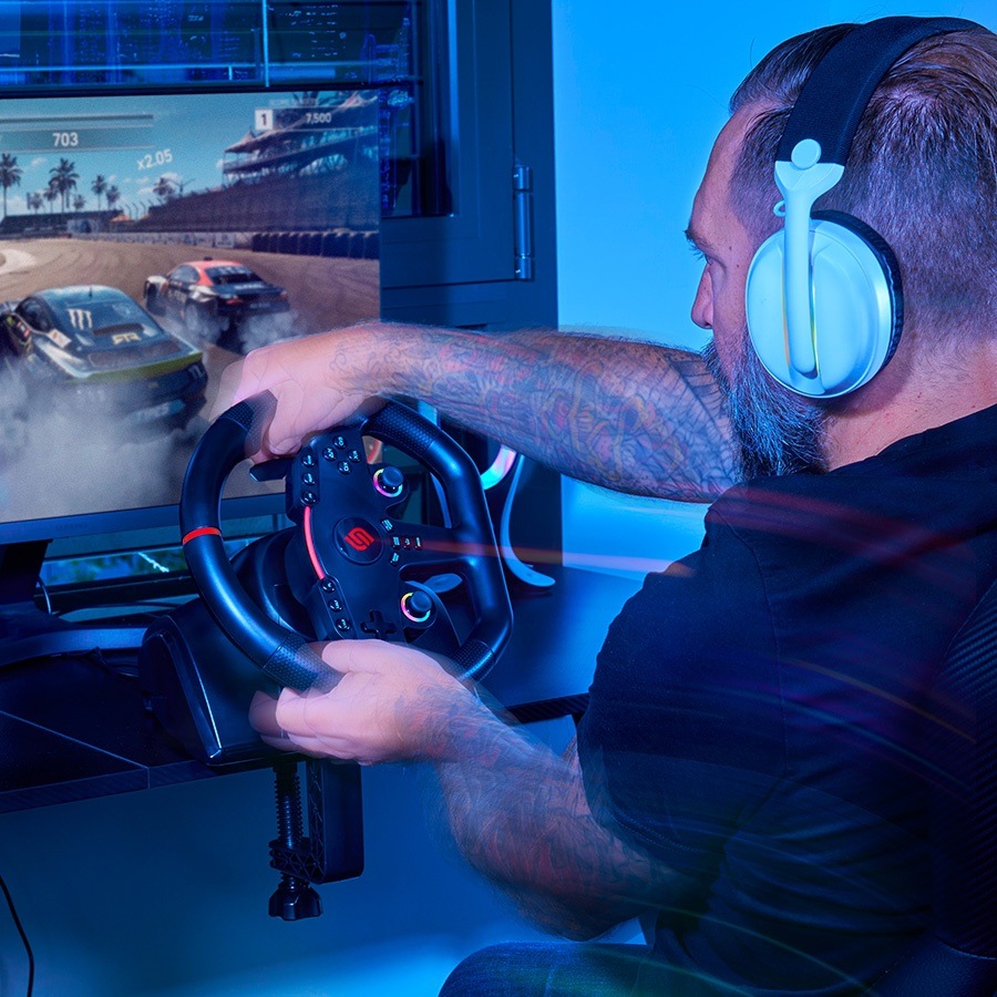Muž so slúchadlami a tetovaním intenzívne hrá závodnú videohru pomocou ovládača na volante s modrým ambientným osvetlením.