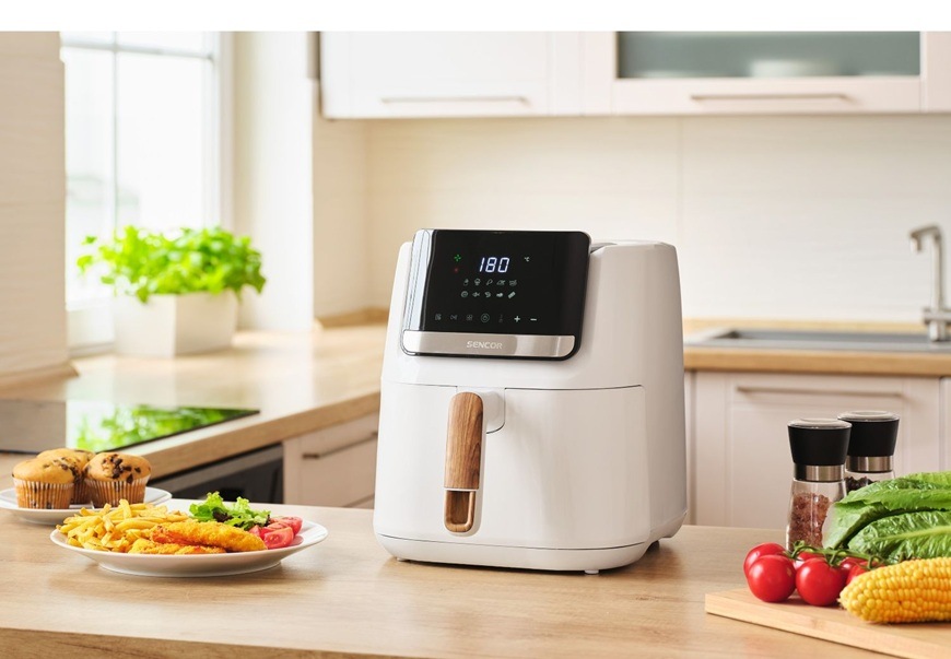 Fritéza teplovzdušná Sencor SFR 6551WH-EUE3 biela Teplovzdušná fritéza Sencor SFR 6551WHEUE3 stojí na svetlej kuchynskej linke a na displeji má nastavenú teplotu 180 stupňov. Vedľa nej leží tanier s hranolkami a vyprážaným jedlom, zatiaľ čo v pozadí modernej kuchyne s oknom a bylinkou pôsobí čisto a sviežo.