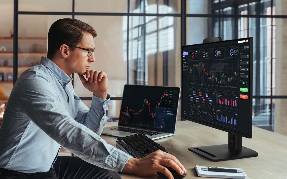 Monitor MSI PRO MP272PMG čierny Muž pracujúci s monitorom MSI PRO MP272PMG a notebookom s finančnými grafmi