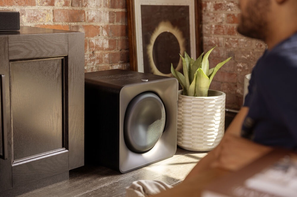 Subwoofer Klipsch Flexus SUB 200 čierny Subwoofer Klipsch Flexus SUB 200 stojí na zemi medzi skrinkou a rastlinou v kvetináči, pred ním sedí človek