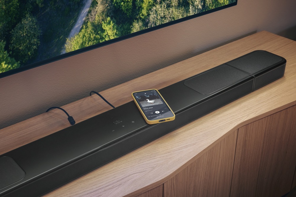 Soundbar JBL BAR 1300 MK2 čierny Soundbar JBL BAR 1300 MK2 položený na televíznom stolíku, na soundbare leží mobilný telefón
