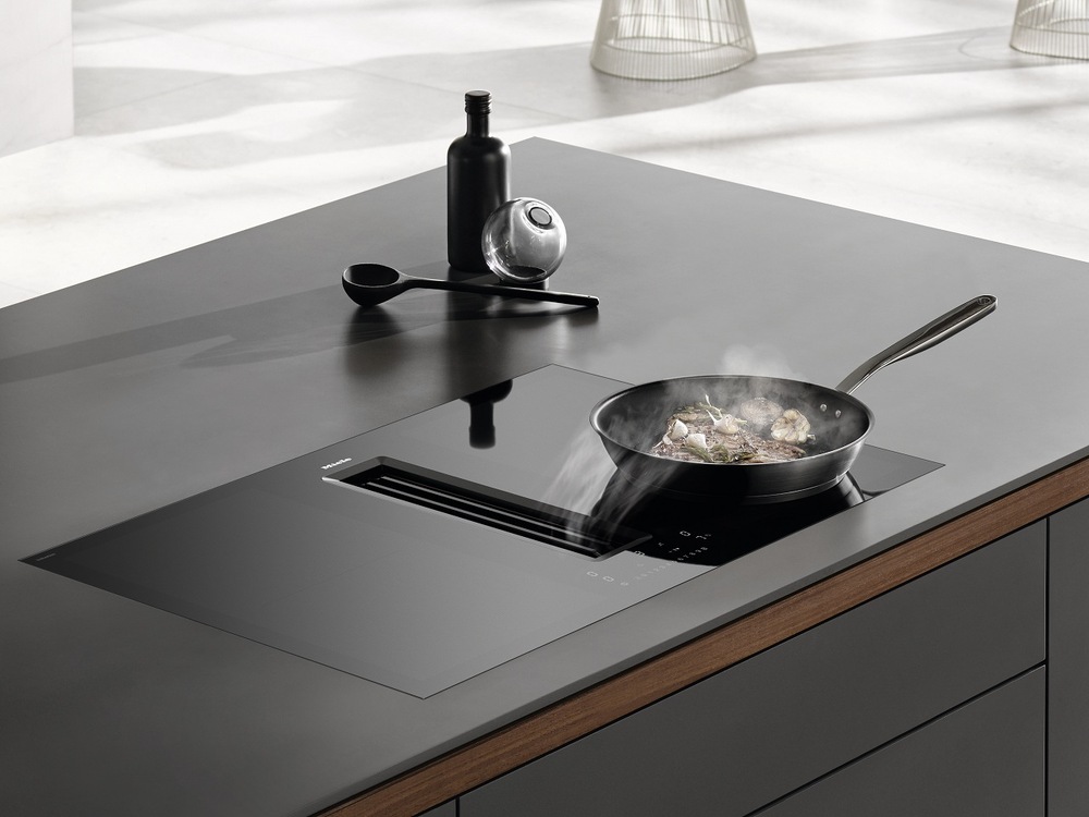 Na indukčnej varnej doske Miele KMDA 7676-1 FL BlackPerfection stojí panvica s pripravovaným jedlom, para z jedla je odsávaná integrovaným odsávačom, vedľa dosky stojí fľaša a leží naberačka