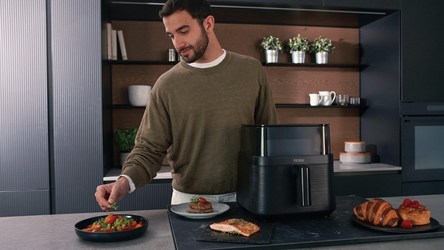 Fritéza teplovzdušná Haier I-Master Series 3 HAF3AFW 011 čierna muž dochucuje jeden z hotových pokrmov, ktoré sa nachádzajú v okolí teplovzdušnej fritézy Haier HAF3AFW 011″ width=