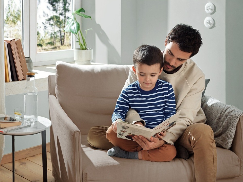 Práčka Electrolux SteamCare® 700 EW7T337DC strieborná Muž sedí s chlapcom v kresle a čítajú spolu knihu. V pozadí je okno do záhrady a pred ním na parapete knihy a izbový kvet. Vľavo vedľa kresla je stolík s fľašou a pohárom vody.