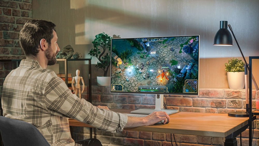 Monitor Lenovo L27h-4A sivý Muž v pruhovanej košeli hrá videohru podobnú League of Legends na monitore v útulnej miestnosti s tehlovými stenami. Teplé osvetlenie a rastliny vytvárajú uvoľnenú atmosféru.
