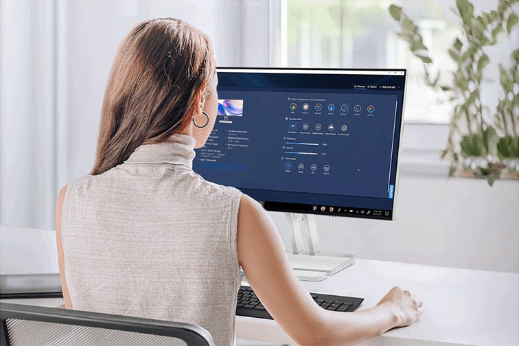 Monitor Lenovo L24-4C sivý Žena pracuje pri stole s notebookom a monitorom, na ktorom sú zobrazené tabuľky a grafy. Svetlá miestnosť s veľkými oknami, rastlinami a pokojnou, sústredenou atmosférou.