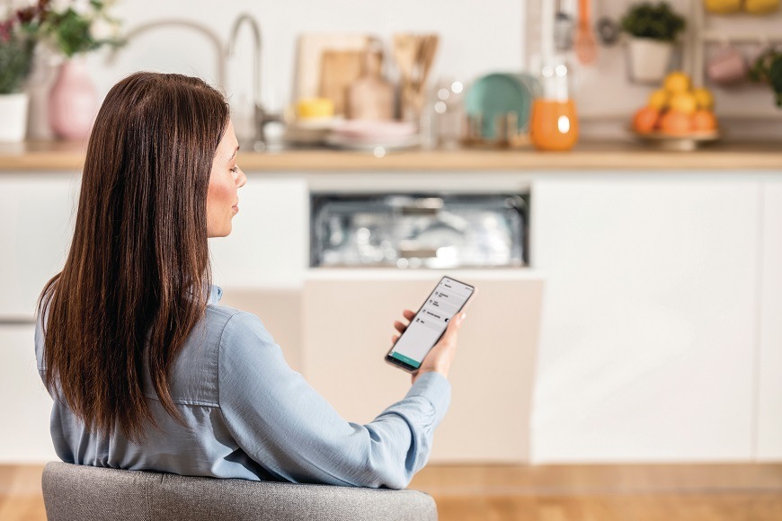 Žena drží v ruke chytrý mobilný telefón av ňom má otvorenú aplikáciu ConnectLife. V pozadí sa nachádza otvorená umývačka Gorenje GVB67365.