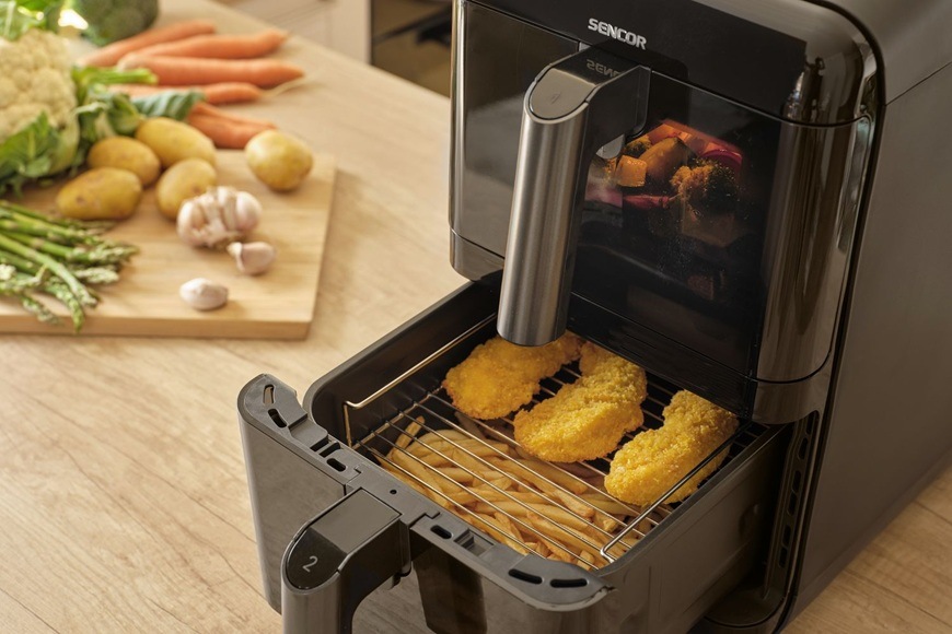 Fritéza teplovzdušná Sencor SFR 1250BK čierna Teplovzdušná fritéza Sencor SFR 1250BK s vysunutým košom, v ktorom sa pripravujú chrumkavé obaľované kúsky mäsa. Na kuchynskej linke vedľa spotrebiča leží čerstvá zelenina ako mrkva, zemiaky a cesnak, čo podčiarkuje domáce varenie a zdravšiu prípravu jedál bez prebytočného oleja.